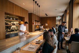 Restaurant ouvert sur la cuisine avec comptoir en béton poli, chef en tenue blanche et espace salle harmonieux en bois chaud